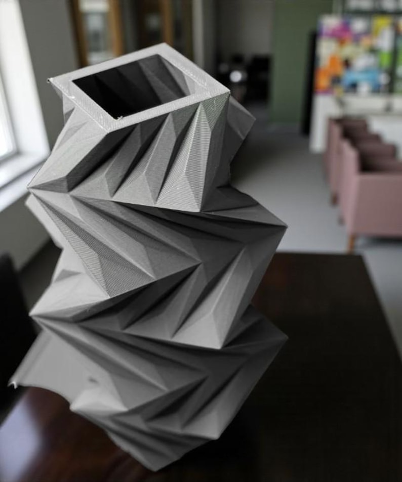 May include: A gray, geometric vase with a square opening and a faceted design. The vase is made of a textured material and sits on a dark surface. The design features sharp angles and a modern aesthetic.