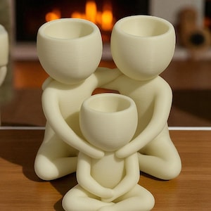 Puede incluir: Una figura familiar de color crema. La escultura representa dos figuras abrazando a una figura m&aacute;s peque&ntilde;a en posici&oacute;n sentada. Cada figura tiene una cabeza redondeada con una abertura en forma de cuenco. La escultura est&aacute; hecha de un material liso y mate.