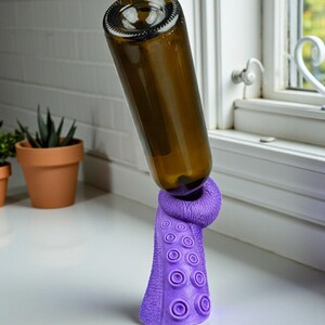 K&ouml;nnte beinhalten: Eine braune Glasflasche wird von einem lila, tintenfischf&ouml;rmigen Weinflaschenhalter gehalten. Der Halter hat ein strukturiertes Design mit kreisf&ouml;rmigen Saugnapfdetails. Die Flasche ist schr&auml;g positioniert. Hintergrund ist eine wei&szlig;e Arbeitsplatte und ein Fenster.