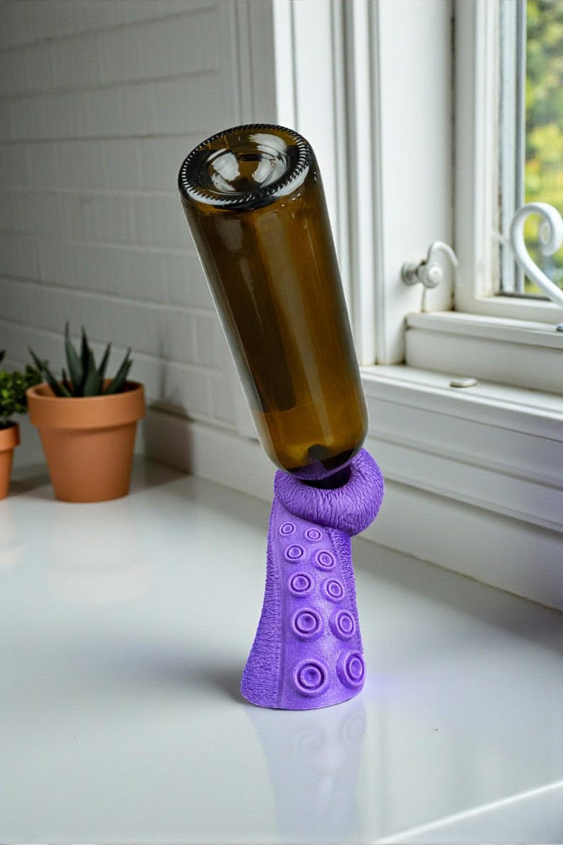 K&ouml;nnte beinhalten: Eine braune Glasflasche wird von einem lila, tintenfischf&ouml;rmigen Weinflaschenhalter gehalten. Der Halter hat ein strukturiertes Design mit kreisf&ouml;rmigen Saugnapfdetails. Die Flasche ist schr&auml;g positioniert. Hintergrund ist eine wei&szlig;e Arbeitsplatte und ein Fenster.
