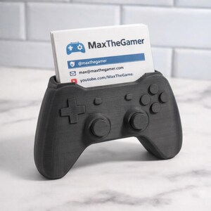 May include: A black business card holder, 3D-printed in the shape of a games controller. The holder contains white business cards with the text "MaxTheGamer" and contact details. The controller features a directional pad, buttons, and analogue sticks.
