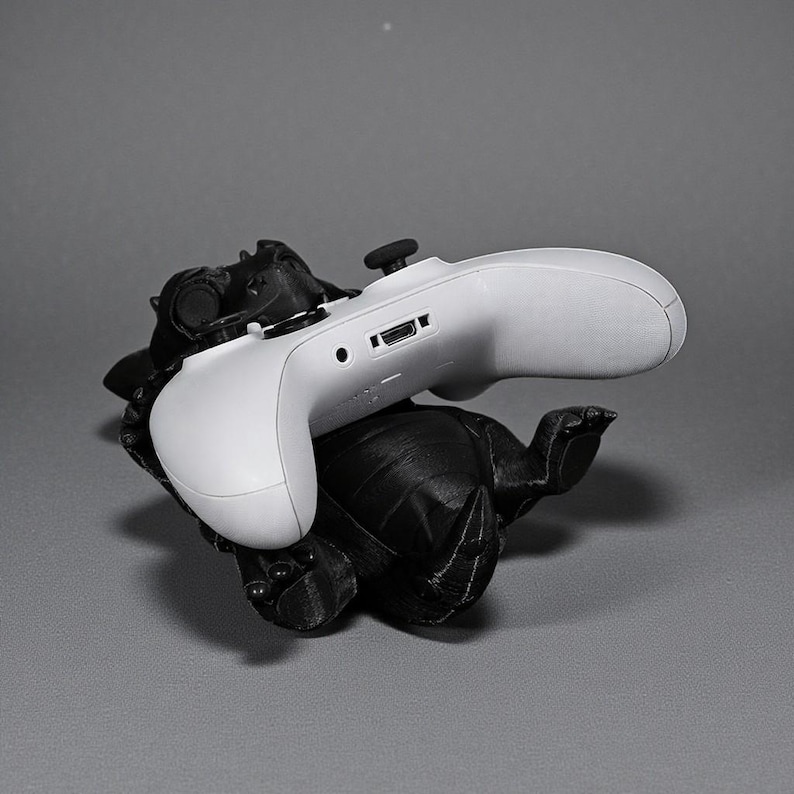 May include: A white Xbox controller sits on a black, turtle-shaped stand. The controller is angled, revealing its buttons and ports. The stand has a textured surface, and the background is a solid grey.