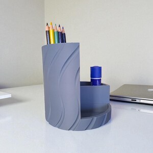 Op de afbeelding: Een grijze bureau-organizer met een golvend ontwerp. Het bevat kleurpotloden en een blauwe lijmstift. De organizer staat op een witte bureau, met een laptop en een potlood op de achtergrond. Een functioneel en stijlvol bureau-accessoire.