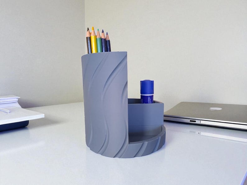 Op de afbeelding: Een grijze bureau-organizer met een golvend ontwerp. Het bevat kleurpotloden en een blauwe lijmstift. De organizer staat op een witte bureau, met een laptop en een potlood op de achtergrond. Een functioneel en stijlvol bureau-accessoire.