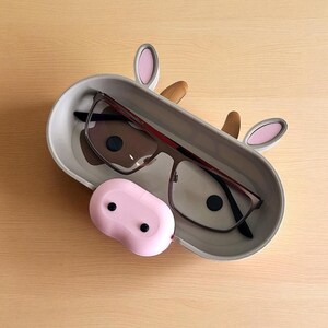 May include: A gray eyeglasses holder shaped like a cow, with pink ears and a brown nose. The holder contains a pair of eyeglasses with a dark frame. A pink, oval-shaped case sits in front of the holder.