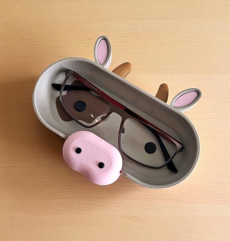 May include: A gray eyeglasses holder shaped like a cow, with pink ears and a brown nose. The holder contains a pair of eyeglasses with a dark frame. A pink, oval-shaped case sits in front of the holder.