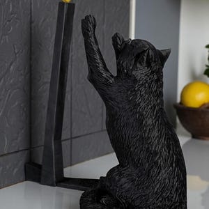 May include: A black cat figurine reaching for a small yellow bird perched atop a black, angular stand. The cat is sculpted with textured fur, and the scene is set against a neutral background, suggesting a decorative item for a home.