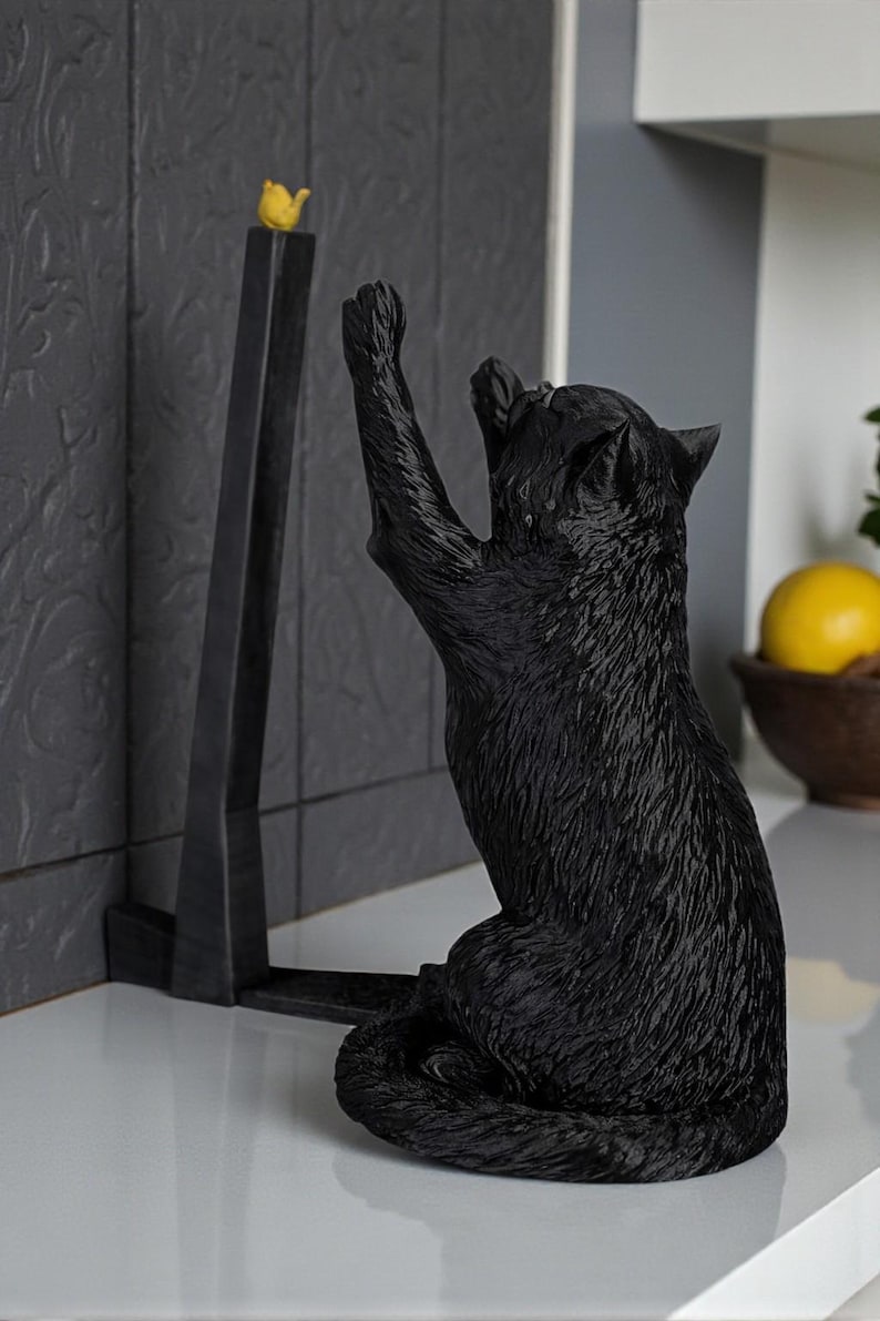 May include: A black cat figurine reaching for a small yellow bird perched atop a black, angular stand. The cat is sculpted with textured fur, and the scene is set against a neutral background, suggesting a decorative item for a home.
