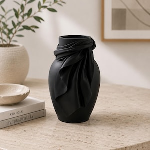 May include: A matte black vase with a draped fabric design around the neck. The vase has a rounded body and a narrow opening. The vase is displayed on a light-colored, textured surface, with a plant and books in the background.