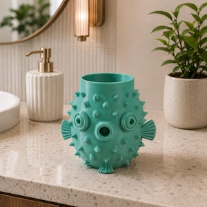 May include: A turquoise, spiky decorative vase resembling a pufferfish. The vase has circular eye-like features and fin-like details. It sits on a speckled countertop, with a white soap dispenser and a potted plant in the background. The vase is a unique home decor item.