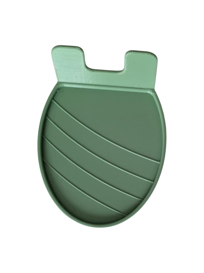 May include: A sage green silicone phone stand. The stand has a rounded base with four horizontal ridges and two rectangular tabs at the top. The stand is designed to hold a phone upright for hands-free use.