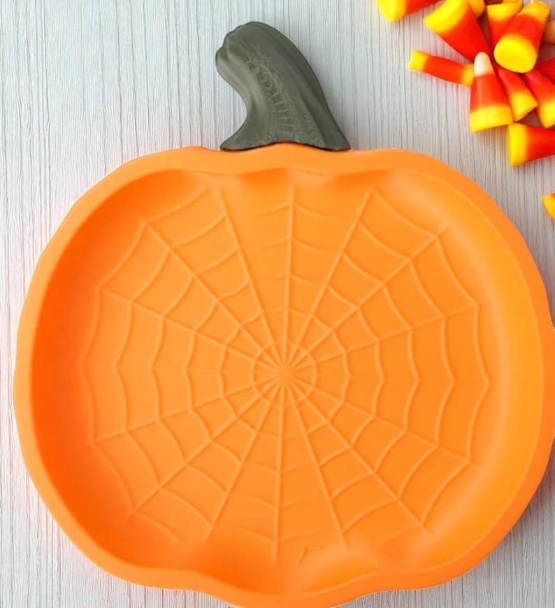 May include: Orange pumpkin-shaped serving dish with a spiderweb design. The dish has a green stem and is filled with candy corn. Perfect for Halloween or fall-themed gatherings.