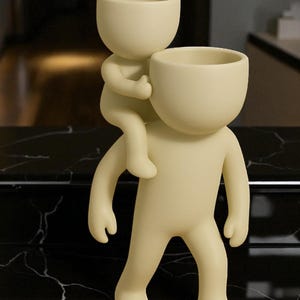 Op de afbeelding: Een cr&egrave;mekleurige decoratieve plantenbak met twee gestileerde menselijke figuren. De ene figuur draagt de andere op zijn rug. Beide figuren hebben open hoofden, wat suggereert dat ze ontworpen zijn om planten of kleine voorwerpen te bevatten. De plantenbak is gemaakt van glad materiaal.