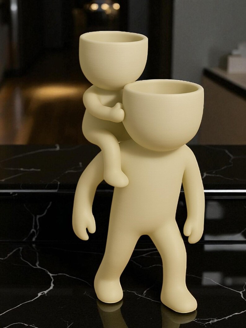 Op de afbeelding: Een cr&egrave;mekleurige decoratieve plantenbak met twee gestileerde menselijke figuren. De ene figuur draagt de andere op zijn rug. Beide figuren hebben open hoofden, wat suggereert dat ze ontworpen zijn om planten of kleine voorwerpen te bevatten. De plantenbak is gemaakt van glad materiaal.