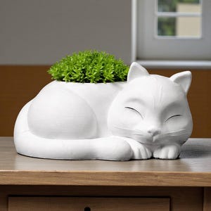 May include: A white ceramic cat-shaped planter, with a sleeping cat design. The planter holds a vibrant green plant. The cat's eyes are closed, and it rests on a wooden table. A drawer with a dark knob is visible below.
