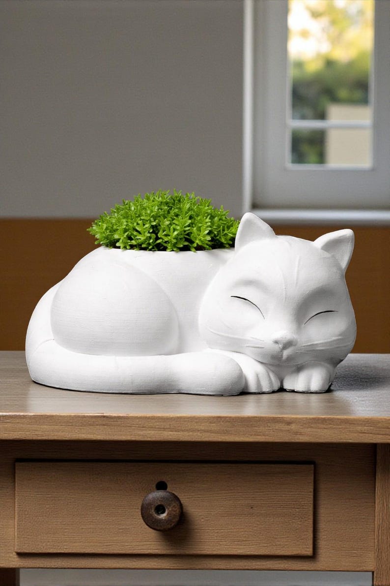 May include: A white ceramic cat-shaped planter, with a sleeping cat design. The planter holds a vibrant green plant. The cat's eyes are closed, and it rests on a wooden table. A drawer with a dark knob is visible below.