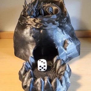 K&ouml;nnte beinhalten: Ein dunkelgrauer, 3D-gedruckter W&uuml;rfelhalter im Drachen-Design. Der Drache liegt in einer h&ouml;hlenartigen Struktur, mit einem wei&szlig;en W&uuml;rfel mit schwarzen Punkten im Vordergrund. F&uuml;r Tabletop-Spiele.