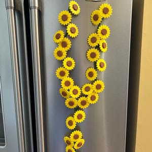 May include: A collection of yellow and brown sunflower magnets arranged in a decorative pattern on a stainless steel refrigerator door. The magnets are in the shape of sunflowers, with yellow petals and brown centers.