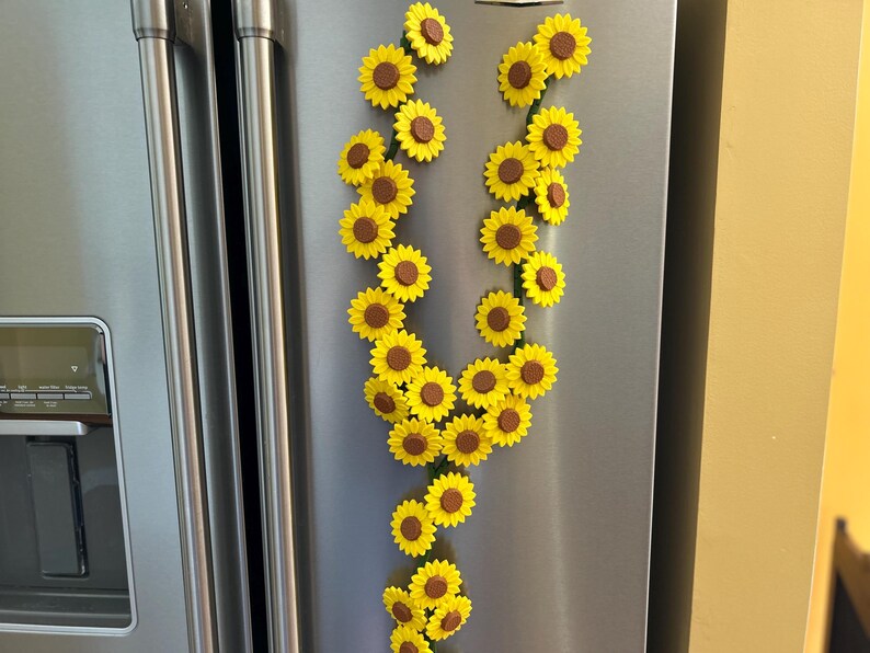 May include: A collection of yellow and brown sunflower magnets arranged in a decorative pattern on a stainless steel refrigerator door. The magnets are in the shape of sunflowers, with yellow petals and brown centers.