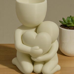 May include: A cream-colored decorative planter in the shape of two figures embracing a heart. The top figure has a cylindrical opening for plants. A small succulent in a white pot is visible in the background.
