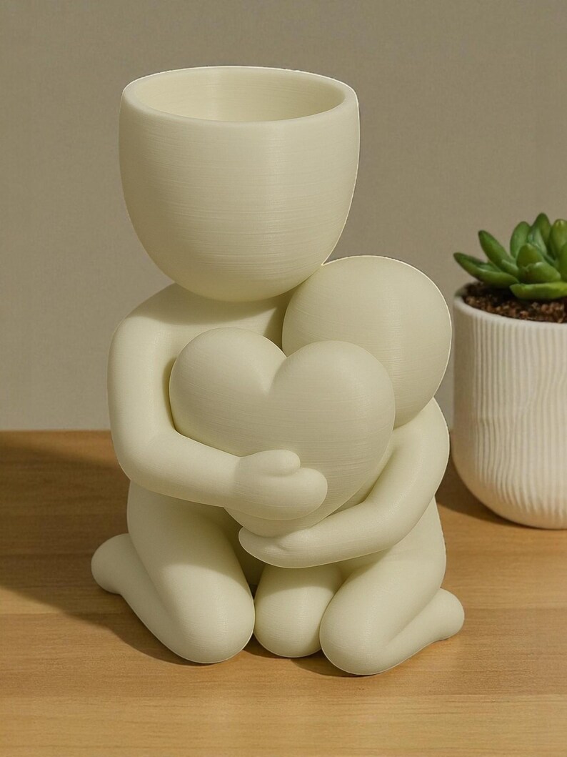 May include: A cream-colored decorative planter in the shape of two figures embracing a heart. The top figure has a cylindrical opening for plants. A small succulent in a white pot is visible in the background.