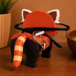 May include: A red panda-shaped eyeglass holder with black legs and a striped tail. The holder has white and brown ears and is holding a pair of black-framed glasses. A silver keyring hangs from the holder.