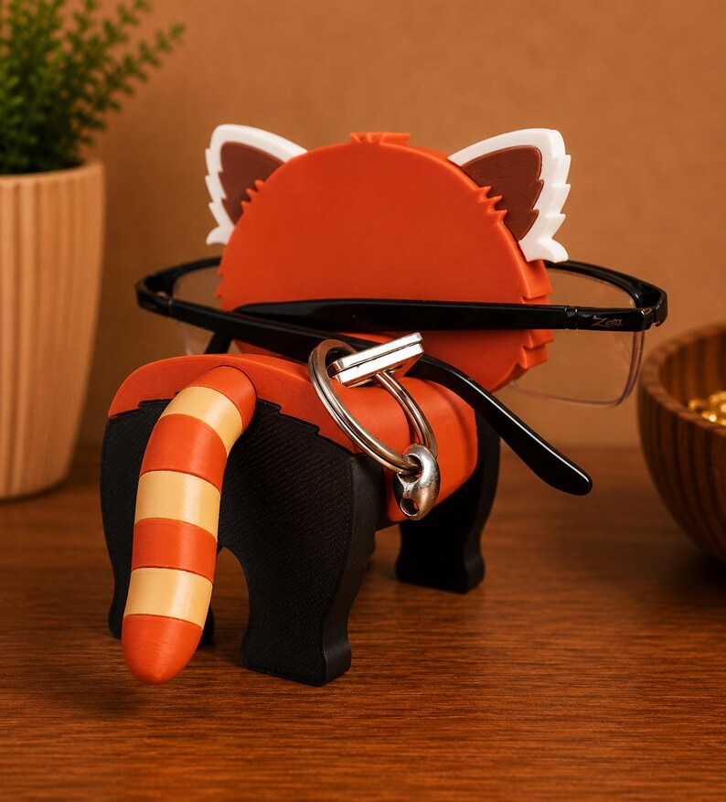 May include: A red panda-shaped eyeglass holder with black legs and a striped tail. The holder has white and brown ears and is holding a pair of black-framed glasses. A silver keyring hangs from the holder.