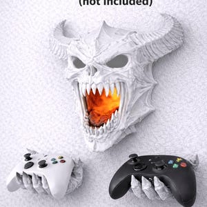 May include: A white, horned demon head wall mount with an open mouth revealing a fiery glow. Two game controller holders, one white and one black, are mounted below. Text reads "mounts to wall with screws (not included)".