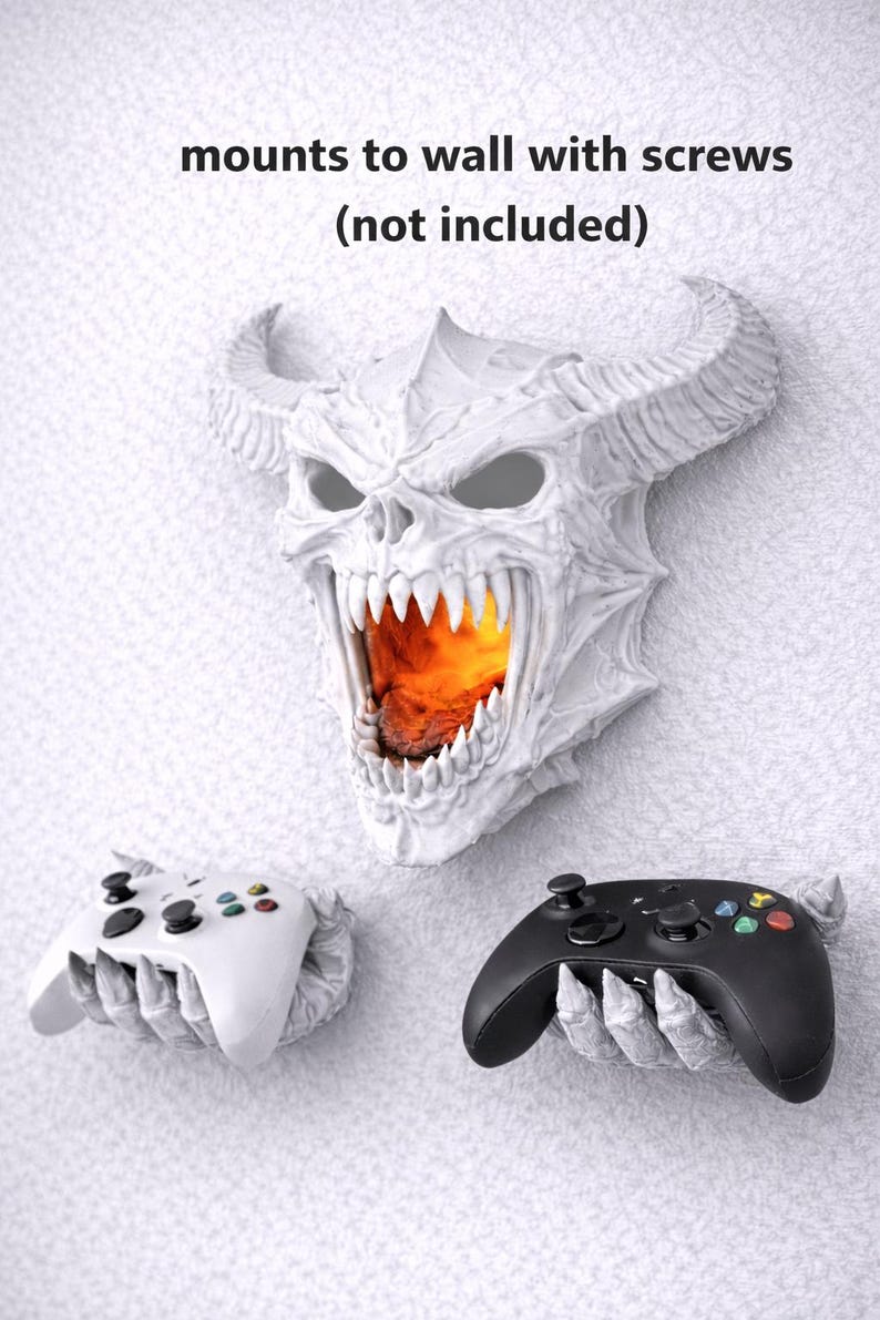 May include: A white, horned demon head wall mount with an open mouth revealing a fiery glow. Two game controller holders, one white and one black, are mounted below. Text reads "mounts to wall with screws (not included)".