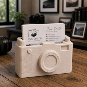May include: A cream-coloured camera-shaped business card holder with a business card that reads "Jane Smith Photography". The business card holder has a lens, viewfinder, and other camera details. The business card has a camera logo with a mountain scene.