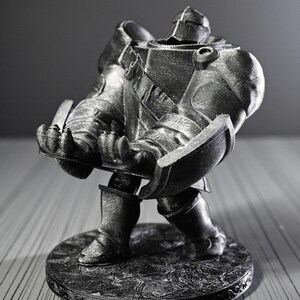 May include: A silver-colored figurine of a knight in full armor, holding a shield. The knight is standing on a round base with an ornate design. The figurine is detailed and appears to be made of metal or a metallic material.