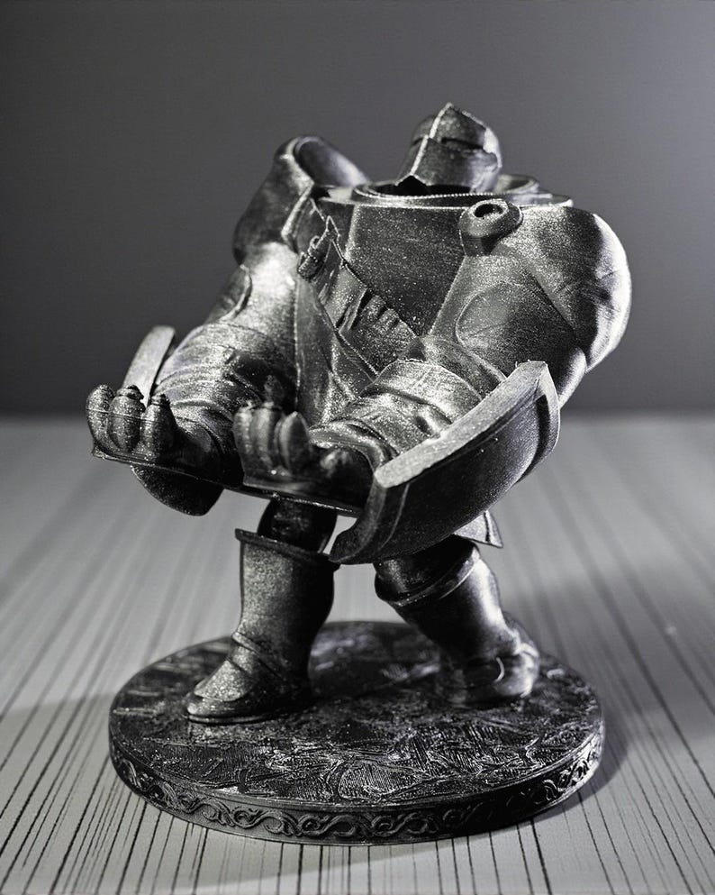 May include: A silver-colored figurine of a knight in full armor, holding a shield. The knight is standing on a round base with an ornate design. The figurine is detailed and appears to be made of metal or a metallic material.