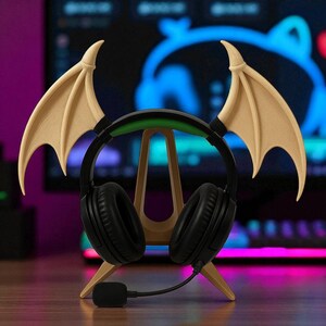 May include: Black gaming headphones with a microphone, resting on a wooden stand. The stand is shaped like a stylized letter A. Attached to the headphones are tan-colored bat wings, adding a unique aesthetic to the setup.