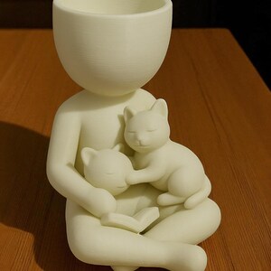 May include: A cream-coloured figurine of a person seated with two cats. The person holds the cats, one in each arm, and is reading a book. The top of the figurine is a planter. The cats are sleeping.