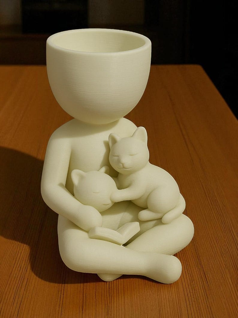 May include: A cream-coloured figurine of a person seated with two cats. The person holds the cats, one in each arm, and is reading a book. The top of the figurine is a planter. The cats are sleeping.