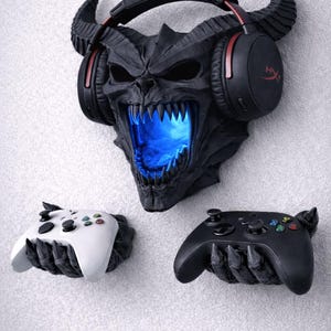 Può includere: Supporto da parete nero a forma di teschio cornuto con bocca blu brillante, che tiene una cuffia nera. Sotto, due supporti per controller, uno bianco e uno nero, ciascuno a forma di mano artigliata. Accessorio per il gaming.