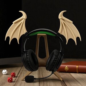 May include: Black headphones with a microphone, displayed on a wooden stand. The headphones feature beige bat wings. Two red dice and a white die are on the wooden surface. A brown book is in the background.