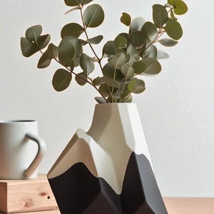Op de afbeelding: Een moderne vaas in de vorm van een bergketen, met een zwart, beige en wit kleurenschema. De vaas bevat groene eucalyptus takken. Een witte mok en een klein houten blok zijn op de achtergrond te zien.