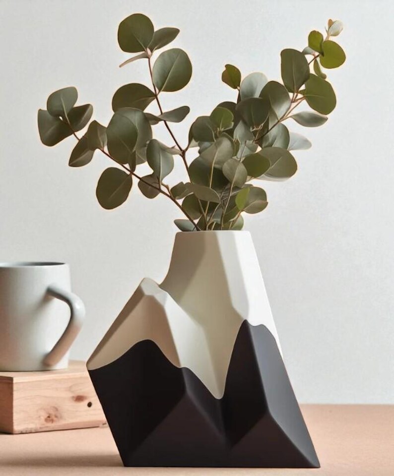 Op de afbeelding: Een moderne vaas in de vorm van een bergketen, met een zwart, beige en wit kleurenschema. De vaas bevat groene eucalyptus takken. Een witte mok en een klein houten blok zijn op de achtergrond te zien.