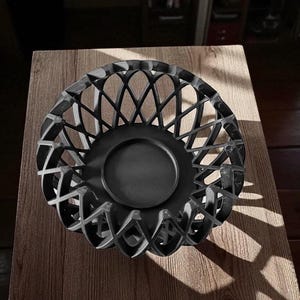 May include: A black decorative bowl with a lattice design. The bowl has a circular base and a detailed, openwork pattern around the sides. The bowl is sitting on a wooden surface.
