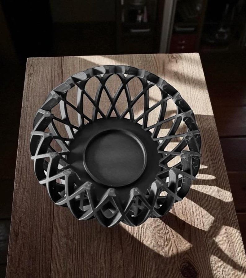 May include: A black decorative bowl with a lattice design. The bowl has a circular base and a detailed, openwork pattern around the sides. The bowl is sitting on a wooden surface.