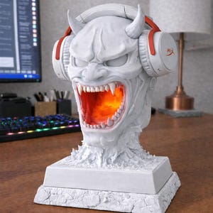 May include: A white, detailed sculpture of a demon head with headphones, lit from within with an orange glow. The headphones have red accents. The text "RGB Puck Light & Remote Included!" is visible. The sculpture sits on a textured base.