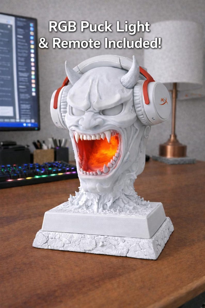 May include: A white, detailed sculpture of a demon head with headphones, lit from within with an orange glow. The headphones have red accents. The text "RGB Puck Light & Remote Included!" is visible. The sculpture sits on a textured base.