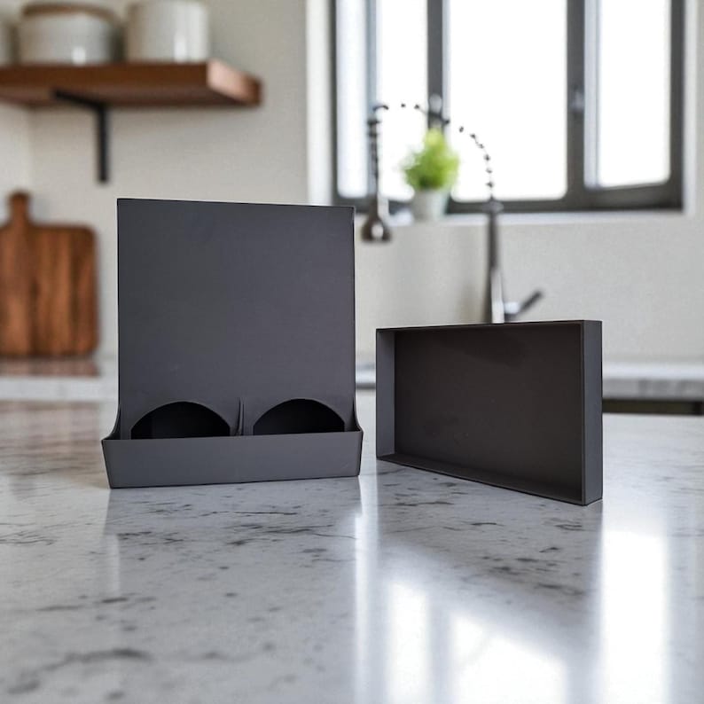 May include: Two dark gray cardboard boxes sit on a marble countertop. One box is rectangular and open, while the other has a vertical back with two circular cutouts. The boxes are likely for storage or display.