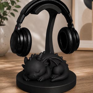 May include: Black headphone stand with a sleeping dragon base. The stand holds a pair of black over-ear headphones. The dragon is detailed with horns, wings, and scales. The stand is on a wooden surface.