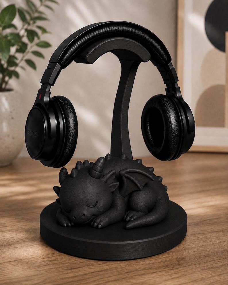 May include: Black headphone stand with a sleeping dragon base. The stand holds a pair of black over-ear headphones. The dragon is detailed with horns, wings, and scales. The stand is on a wooden surface.