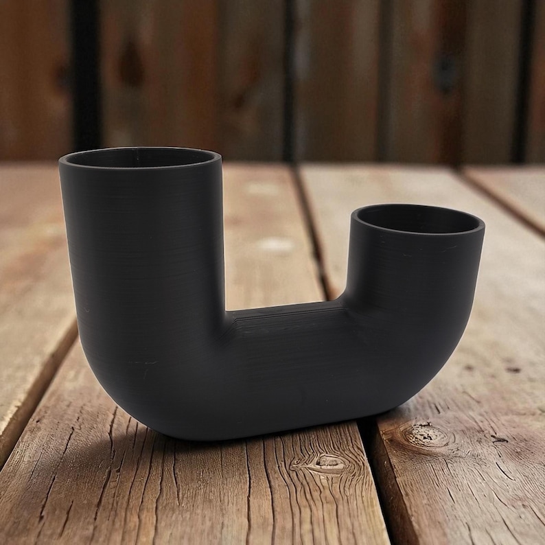 May include: A matte black, U-shaped ceramic vase with two openings. The vase sits on a wooden surface. The design is modern and minimalist, suitable for displaying flowers or as a decorative accent.