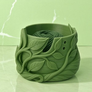 May include: A green yarn bowl with a leaf design. The bowl is filled with a ball of green yarn. The bowl has two holes on the side and is made of a solid green material.