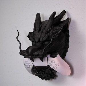 May include: A black dragon head wall mount holding a white and black video game controller. The dragon has detailed features and a textured surface. The controller has a symmetrical design with buttons and joysticks.