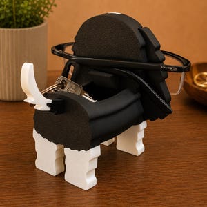 Pu&ograve; includere: Un portachiavi e portaocchiali decorativo a forma di cane bianco e nero. Il supporto ha una fessura per gli occhiali e un portachiavi. Il supporto &egrave; realizzato in schiuma e si trova su una superficie di legno.